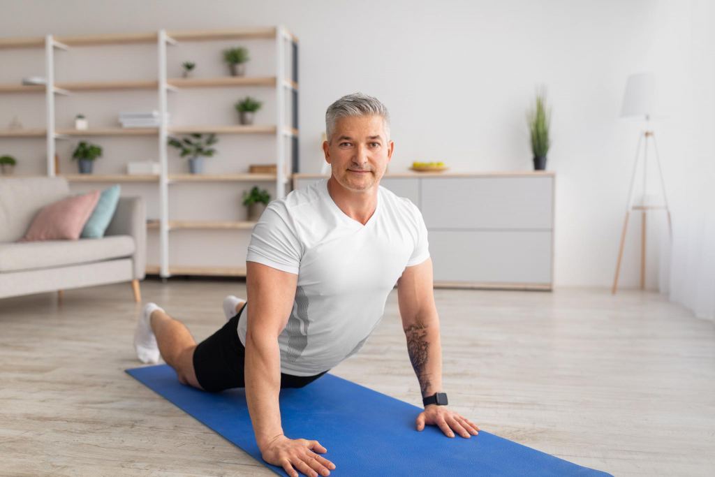 Mature man stretching back muscles, doing sphinx cobra pose or upward-facing dog asana, excercising in living room interior on yoga mat, empty space