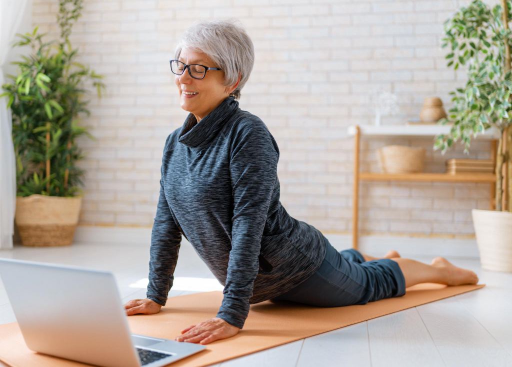 Senior woman in activewear watching online courses on laptop while exercising at home.
